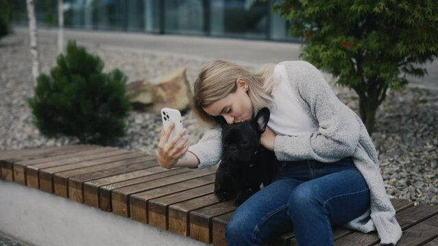 Beautiful Smiled Woman is Sitting on the Bench with Her Lovely Black French Bulldog and Doing Selfie, Making Memories with Her Puppy. Technology and Dogs Friendship Concept
