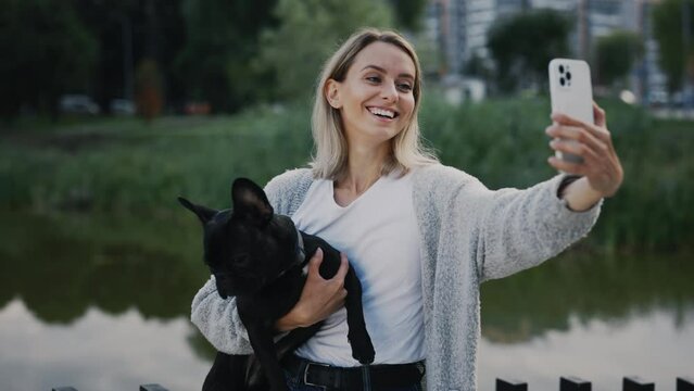 Beautiful Happy Woman Is Standing near the Local Lake with Her Lovely Black French Bulldog and Doing Selfie, Making Memories with Her Cute Puppy. Technology and Dogs Friendship Concept