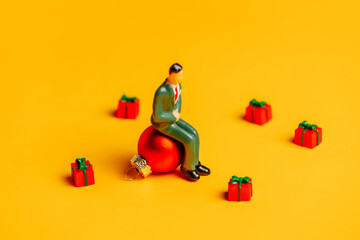 Miniature figure of man sits on Christmas bauble surrounded by gifts on yellow background
