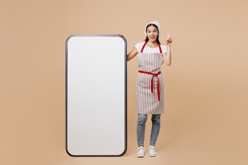 Full body young housewife housekeeper chef baker woman wears apron toque hat big huge blank screen mobile cell phone smartphone point finger up isolated on plain beige background. Cook food concept.