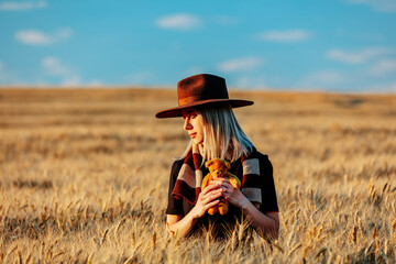 Blonde woman in black dress and hat with scarf holds teddy bear toy in wheat field in autumn season time