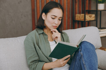 Obraz premium Young calm serious minded smart woman of Asian ethnicity wearing casual clothes read book novel sits on grey sofa couch stay at home hotel flat rest relax spend free spare time in living room indoor.