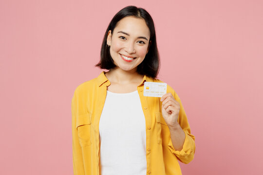 Young Smiling Happy Woman Of Asian Ethnicity Wear Yellow Shirt White T-shirt Hold In Hand Mock Up Of Credit Bank Card Isolated On Plain Pastel Light Pink Background Studio Portrait. Lifestyle Concept.