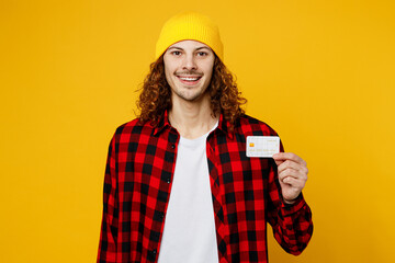 Young siling cheerful cool caucasian man wearing red checkered shirt white t-shirt hat hold in hand mock up of credit bank card isolated on plain yellow background studio portrait. Lifestyle concept.