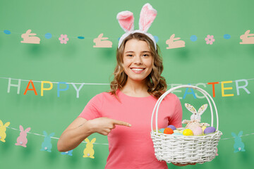 Young woman wear pink casual clothes bunny rabbit ears hold point index finger wicker basket with eggs isolated on plain pastel light green background studio portrait Lifestyle Happy Easter concept