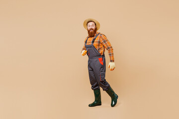Full body young fun happy bearded man wears straw hat overalls work in garden walking going looking camera isolated on plain pastel light beige color background studio portrait. Plant caring concept.