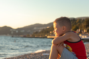 Cute kid  boy sitting on the beach near sea and looking on the waves, boy on the seaside in sunset.