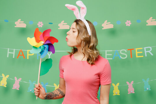 Young smiling happy cheerful woman wear pink casual clothes bunny rabbit ears blowing on toy windmill isolated on plain pastel light green background studio portrait. Lifestyle Happy Easter concept.
