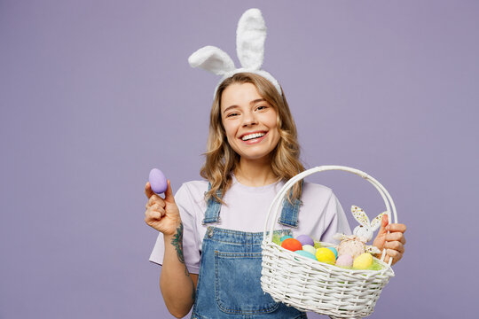 Young smiling positive fun cheerful woman wearing casual clothes bunny rabbit ears hold wicker basket show colorful egg isolated on plain pastel purple background studio portrait Happy Easter concept