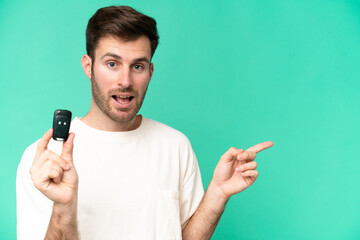 Young caucasian man holding car keys isolated on green background surprised and pointing finger to the side