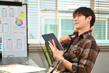 Thoughtful asian man UX graphic designer sitting at creative workplace and contemplating new...