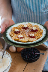  A lady in a blue striped apron holds a plate with a waffle garnished with strawberry jam and cream.