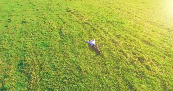 Aerial view point. Low orbital flight around man lied on green grass with notebook (pad) at yellow rural field. Casual weared. Sun rays. Radial movement.