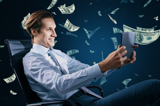 Digital business, success, money, freedom and power concept with happy businessman on office chair using digital tablet on dark background under dollar banknotes rain