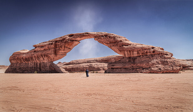 A "Dust Devil" or small tornado passes through "Rainbow Rock", also known as "Arch Rock' in Alula, Saudi Arabia. A man wearing traditional Clothes stands right under the arch.