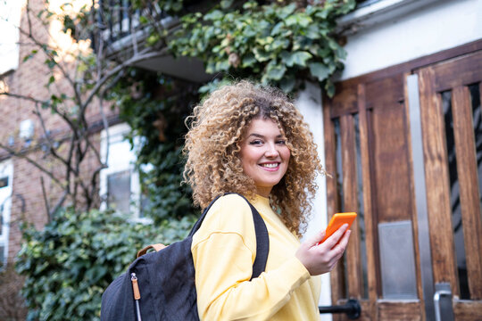 Smiling Young Woman With Curly Hair Holding Smart Phone Outside House