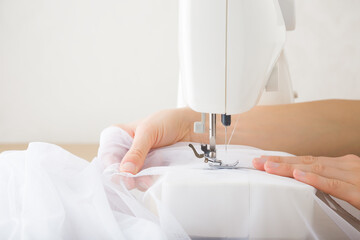 Young adult woman hands working with sewing machine and sewing white day curtains. Closeup. Side...