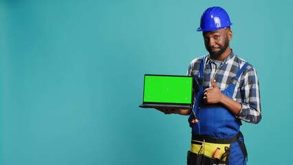 Happy mechanic pointing at laptop with greenscreen on camera, advertising isolated template display on pc. Young construction worker showing computer with chroma key mockup copyspace.