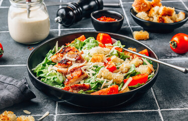 Homemade Caesar salad with crispy chicken and tomatoes on a black tiled background