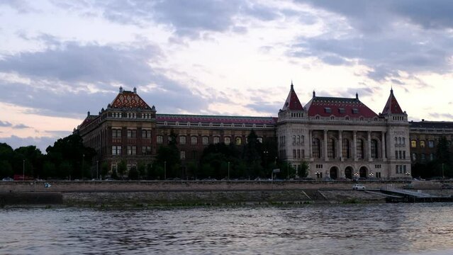 Cinematic shot of Budapest University of Technology and Economics BME building at golden hour, shot from the Danube River in Budapest, Hungary. Evening clouds in the sky, street lights glow. 4k 30fps