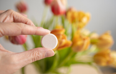 round large tablet, vitamin in the girl's hand, against the background of yellow tulips, indoors. Flu and cold season, new virus strain. Lack of vitamins in spring and winter, uncontrolled medication