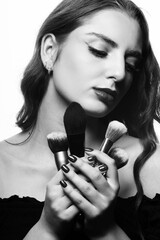 Makeup and fashion concept. Portrait of beautiful woman with wavy hair holding various make-up brushes near her face. Model with closed eyes looking aside the camera. Black and white image