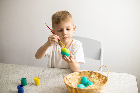 A child sitting at a table paints eggs with a brush. Nearby are paints and a wicker basket. Easter