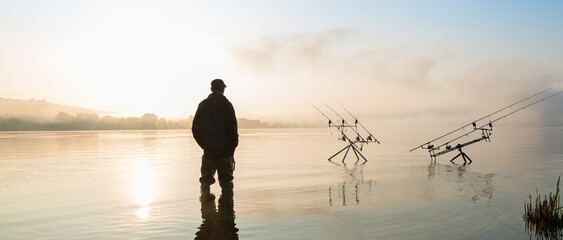 Fishing adventures, carp fishing. Fisherman with boots on the lake at sunrise on a misty morning