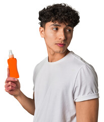 A young latin man holding a solar protection cream looks aside smiling, cheerful and pleasant.