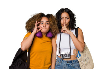 Two diverse friends on a beach vacation with vintage camera, beach bag, and travel pillow with fingers on lips keeping a secret.