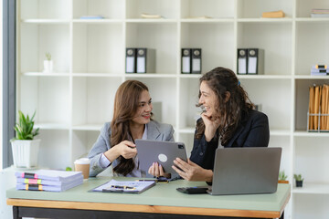 Two young cheerful businesswoman working using digital tablet at conference table with work papers at office. Female business colleague using touch screen computer for real estate project discussion