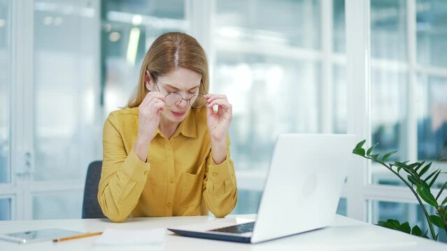 Exhausted stressed blond mature business woman takes off glasses to massage eyes with pain ache hurt bad poor vision after computer overwork at glass workplace Visual fatigue and eyestrain concept