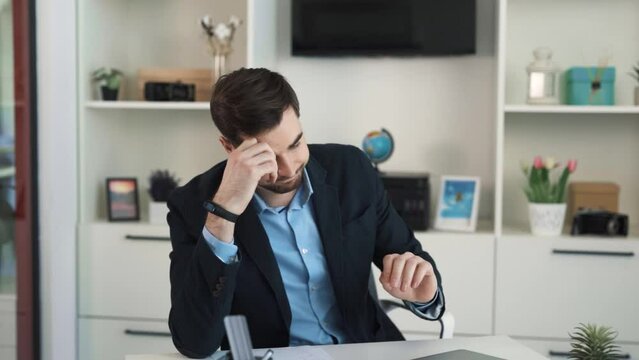 Confused and worried man sitting at desk in bright office, bored without work and looking at clock