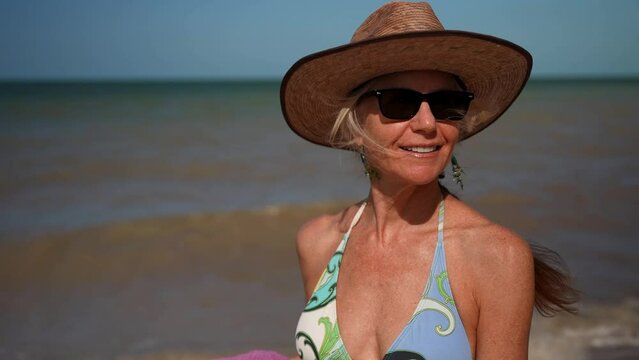 Extreme closeup of happy smiling mature senior woman 50s, 60s, in bikini straw hat sunglasses, walking on the beach 