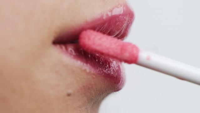 Woman uses soft applicator to apply bright pink gloss on lips on white background. Young female person shows makeup process extreme closeup