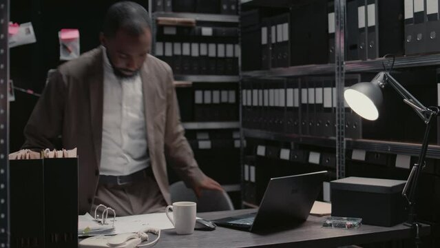 African american agent reading suspicious files in archive room, trying to solve crime and catch suspect. Private inspector working with detective map and forensic evidence. Handheld shot.