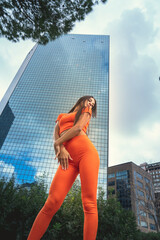 Photo shoot of stylish caucasian hip hop dancer posing having fun in orange leggings and top gymnastic jumpsuit, enjoy the urban town as background. City buildings shooting of moving fitness model