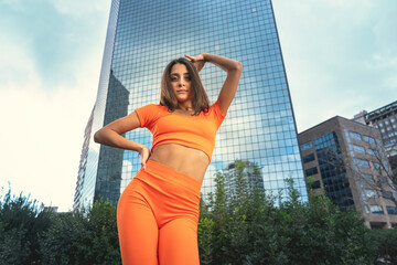 Photo shoot of stylish caucasian hip hop dancer posing having fun in orange leggings and top gymnastic jumpsuit, enjoy the urban town as background. City buildings shooting of moving fitness model