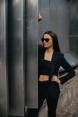 Fashion portrait of young elegant woman outdoors. Black suit, sunglasses, gray metal background.