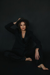 Portrait of a modern stylish woman. A brunette in a suit and hat poses barefoot on a black...