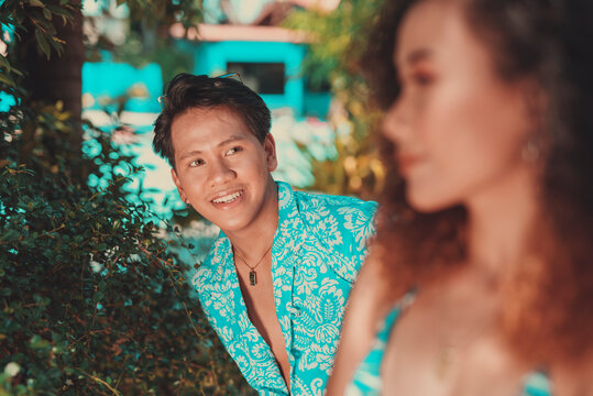 Asian Man wearing unbuttoned hawaiian luau polo shirt smiling at a woman in foreground. Male and female having fun outside.