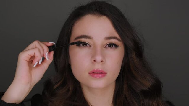 Young woman applies black mascara on long eyelashes standing on dark background. Blogger does makeup looking in camera closeup slow motion