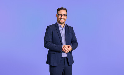 Smiling elegant mid adult male manager dressed in blue suit clasping his hands while standing confidently isolated over blue background - obrazy, fototapety, plakaty