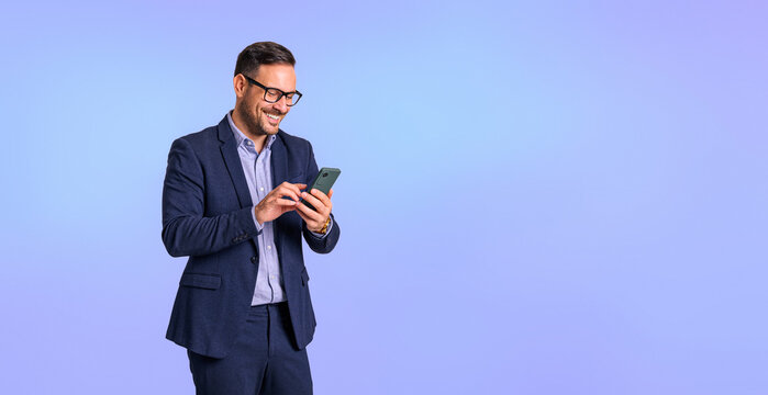 Smiling mid adult businessman dressed in elegant formalwear texting online over smart phone and standing isolated on blue background