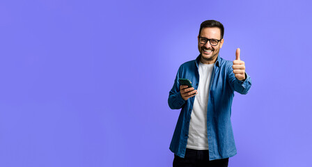 Portrait of cheerful young adult man in denim shirt using smart phone and showing thumbs up sign...
