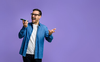 Excited young adult man screaming cheerfully while talking over smart phone speaker and standing isolated against purple background