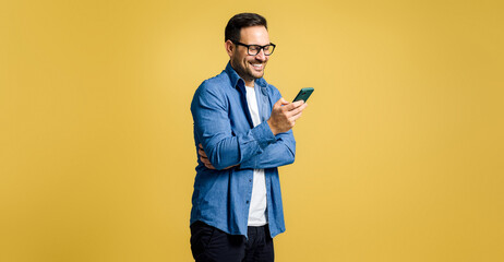 Smiling handsome young adult man dressed in denim shirt chatting over smart phone and standing isolated against yellow background