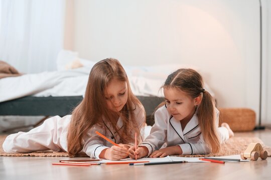 Two little girls are lying down on the ground and drawing together in domestic room