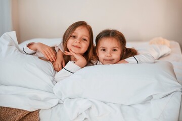 Smiling, laying down on the bed. Two little girls are playing and having fun together in domestic room