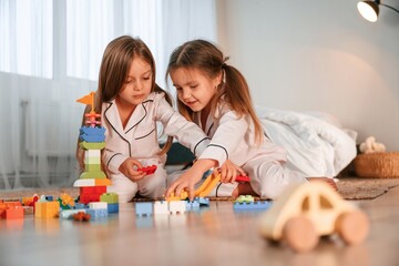 Bunch of plastic construction toys are on the floor. Two little girls are playing and having fun together in domestic room